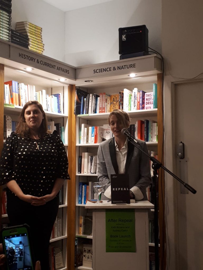 Sydney Calkin (L) and Kath Browne (R) at the launch on 31 January 2020 of their edited book, After Repeal. Kath Browne is at the podium, microphone in front of her. Behind them both are shelves of books.