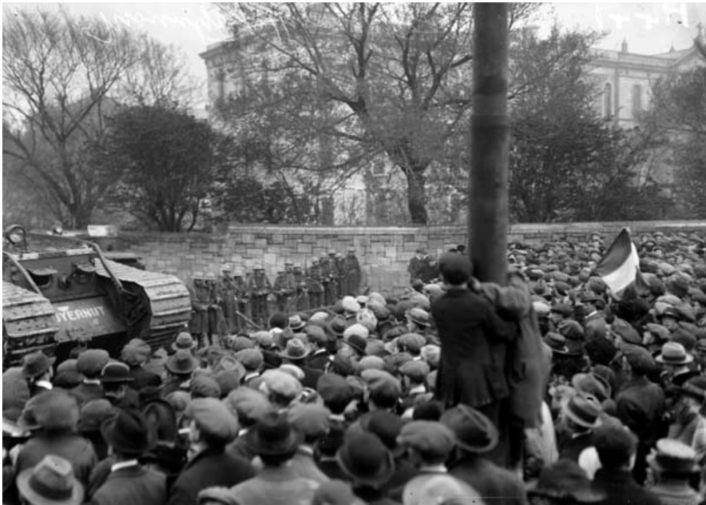 A black and white photograph taken during Ireland's biggest general strike. Against a background of the Mountjoy Prison a crowd on the right is faced off against a line of British soldiers and tank on the left. The crowd are mostly men but there are many women, all are wearing caps and hats. Two boys have climbed a pole to get a better view.