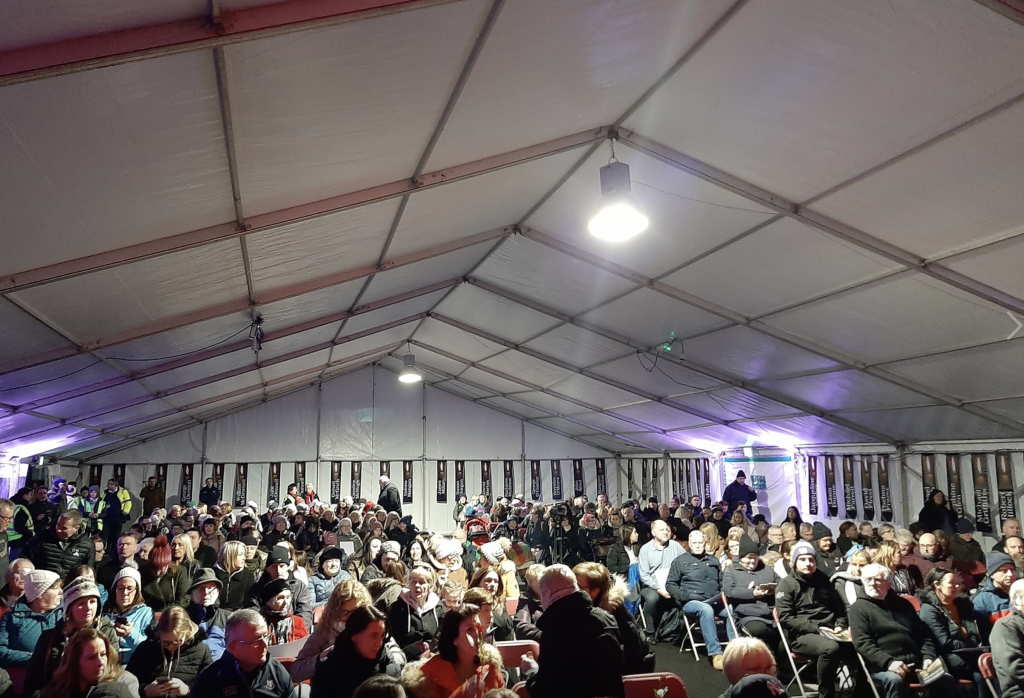 A full marquee at the moving Stardust 48 memorial, 13. February 2020. The picture shows a marquee with temporary lighting and chairs, It is full with people standing at the back.