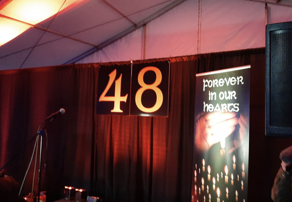 The stage at the marquee for the 13 February 2020 memorial event for the Stardust 48. It shows a microphone, a red curtain and in front of it in large yellow figures: 48. A pop-up banner on the right of the pictures says: forever in our hearts.