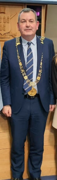 Tom Brabazon in a blue suit, wearing the Mayoral chains of office for Dublin City and while wearing a smile, looking a little uneasy, with clenched right fist and hunched shoulders.