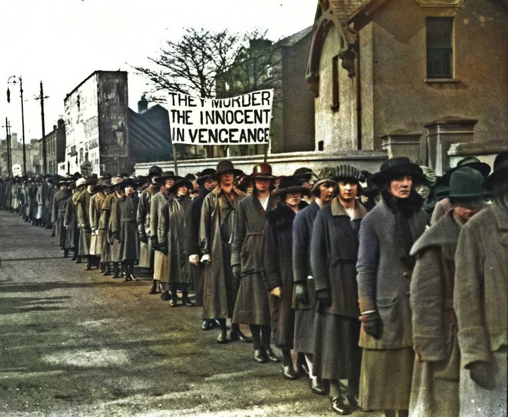 Colourised newspaper image of women in a long row, wearing coats and dark skirts. They are marching along a north Dublin street towards Mountjoy Jail and in the middle of the picture are women carrying a banner saying: They Murder the Innocent in Vengeance.