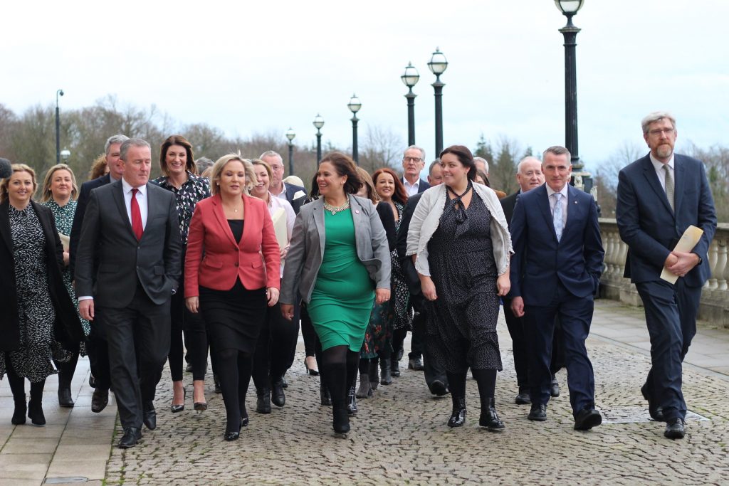December 2019 and Sinn Féin leader Mary Lou McDonald alongside Deputy First Minister Michelle O'Neill pose with the newly appointed Ministers and Sinn Féin members of the Northern Ireland Assembly. They are walking on cobbled stones, all in suits, McDonald in a Green dress, O'Neill in a red jacket.