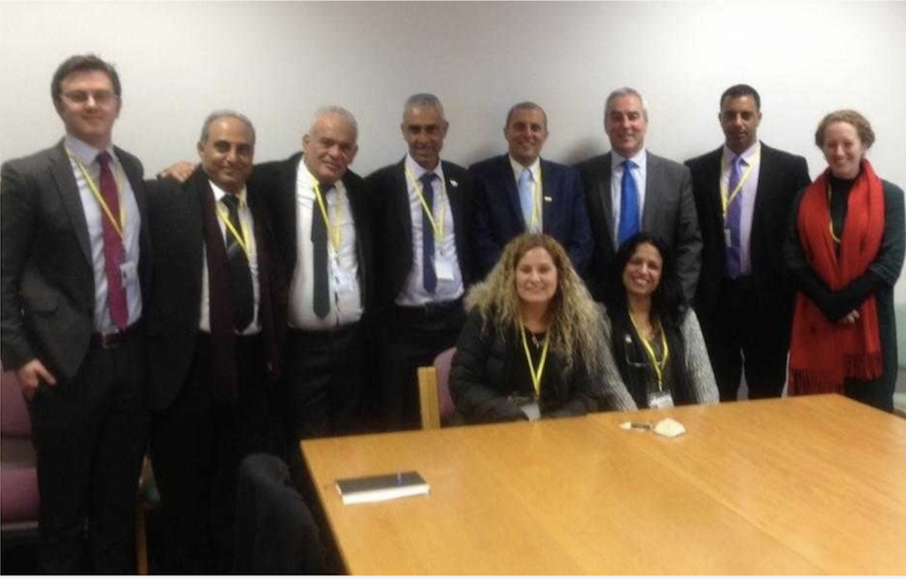 In June 2019 a delegation from Israel's Likud party visiting Belfast where they met with Sinn Féin members, including Pat Sheehan. Seven men with suits and ties stand at the back, one woman standing on the far right. Two women are seated at a polished wooden stable in front of the standing members of the delegation.