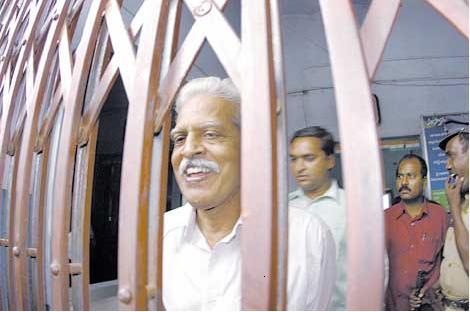 A smiling poet, Marxist and Telangana activist Varavara Rao looks out from behind shutters. Rao has been arrested multiple times by the Indian state but his most recent imprisonment has led to his contracting COVID19.